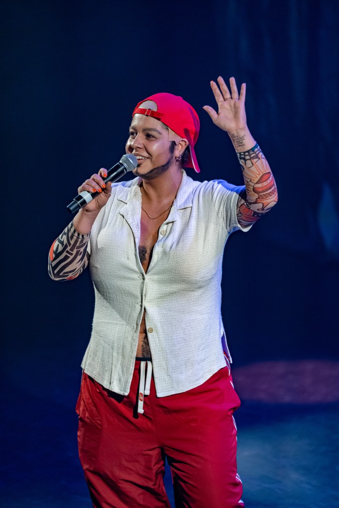 Nati Daddy is waving to the audience with fake tattoos, Red cap on backwards, white shirt and red trackpants, talking into a microphone. The background is a dark blue/purple haze.