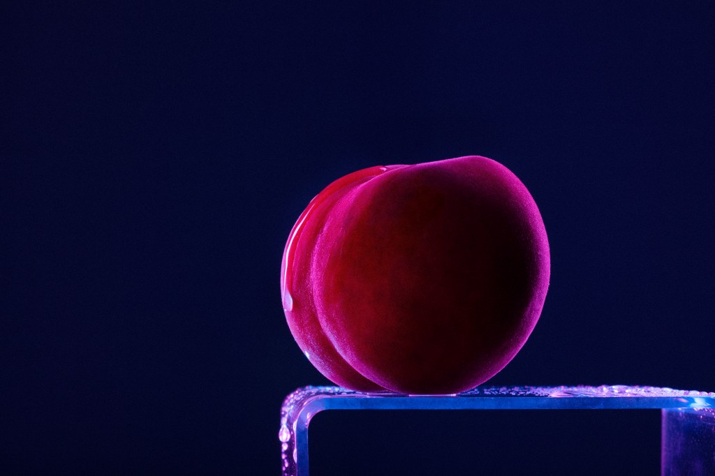 White, Thick, Sans Serif text reads 'Big Thick Energy Taking up Space' It's overlaid on an image of a peach sitting on a table. The peach has one oozing dribble down its left side. It's lit with purple or pink lighting and is on a navy background.