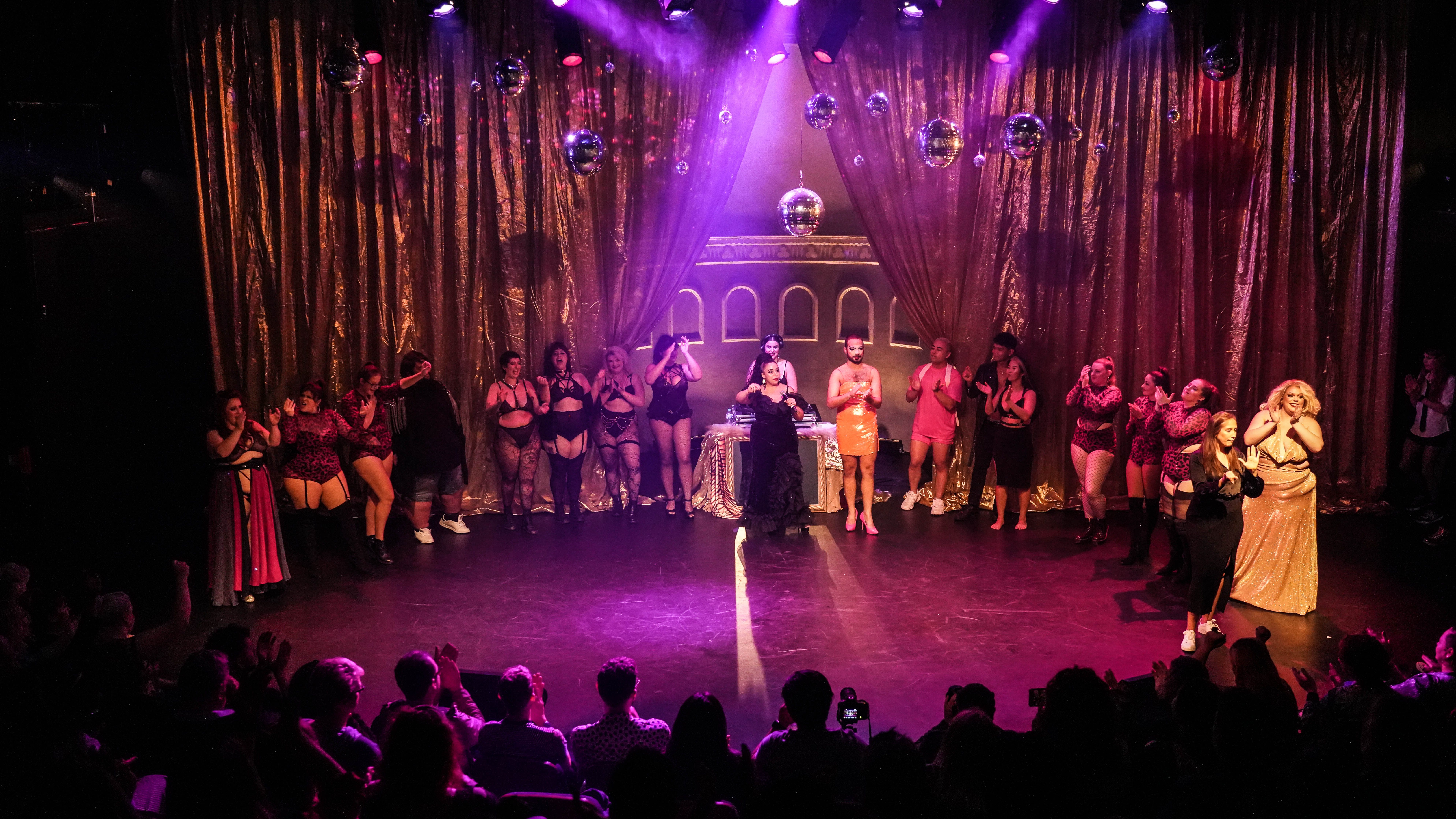 The cast of Big Thick Energy's March Finale standing on stage, forming a circle with the front row of the audience. Demon Derriere is wearing a black dress and holding a microphone in the centre of the image, standing next to Peach Fuzz.  The people on stage are all clapping, cheering, laughing or dancing. There is gold sparkly curtains in the background and different sized disco balls hanging above everyone reflecting purple and pink light. The audience is clapping. 